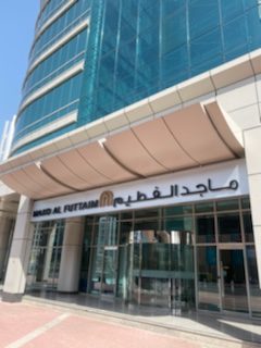 Glass skyscraper revolving door entrance at street level on a sunny day; English and Arabic company sign